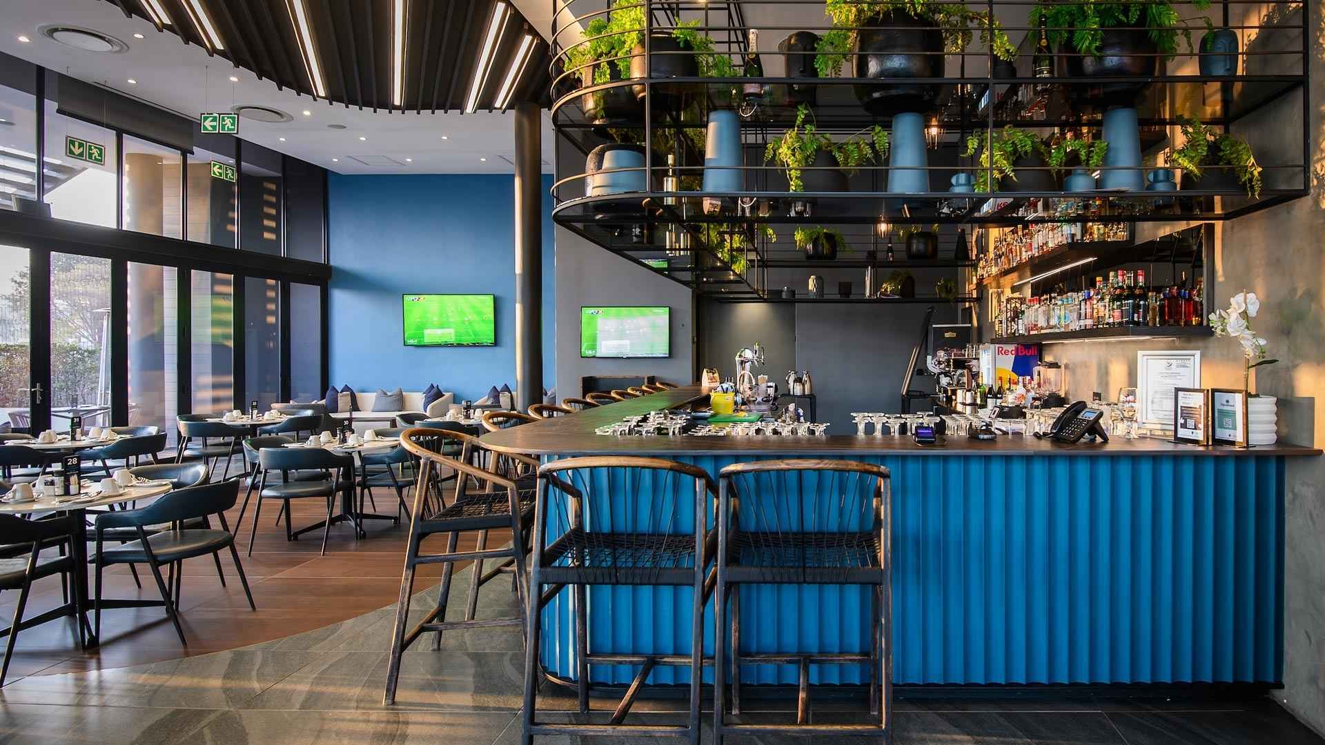 Hotel bar with a blue counter, bar stools and plant-filled shelves